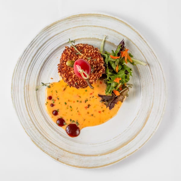 A crispy soul-food patty with a red pepper garnish, green salad, and creamy orange sauce on a white plate.