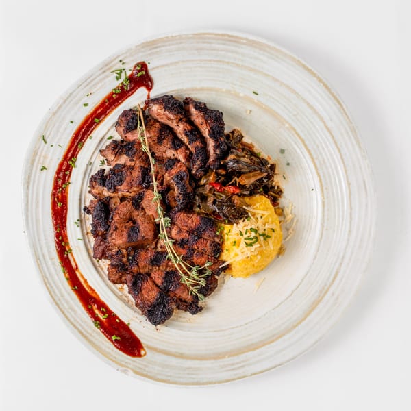 Grilled steak slices with collard greens, cheesy grits, and a red sauce drizzle on a white plate.