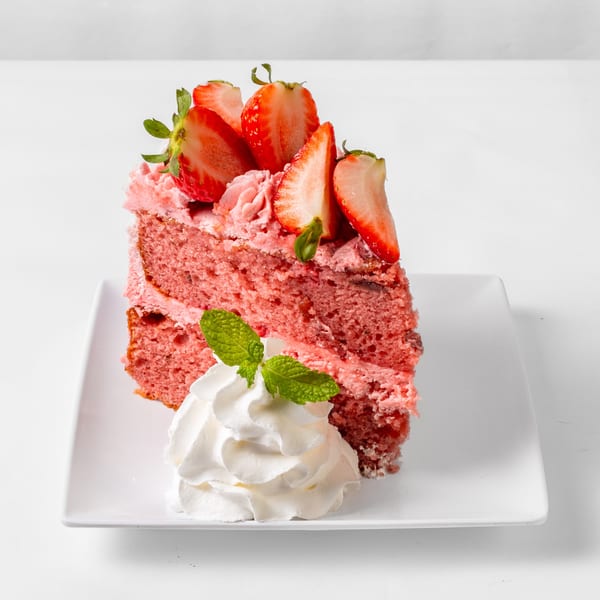 A slice of strawberry cake with pink frosting, fresh strawberries, whipped cream, and mint.